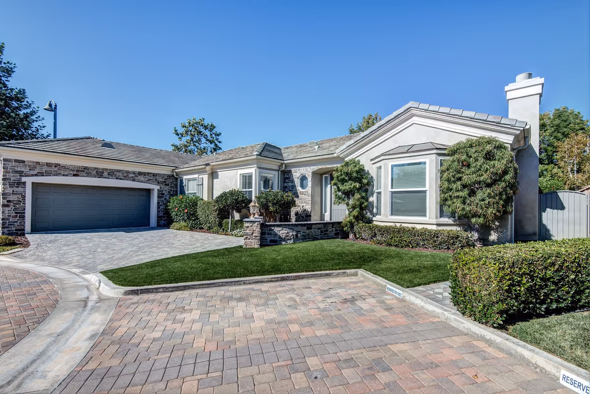Single-story residential building with an attached garage, paved driveway, and manicured lawn and shrubs under a clear blue sky.