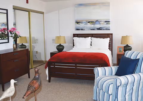 A cozy bedroom featuring a wooden bed with white pillows and a red blanket, flanked by two green lamps on dark wooden nightstands. A striped armchair with a blue cushion is positioned in the foreground, and a wooden dresser with a vase of pink flowers and a mirror is on the left. The room has beige carpeting, a large painting above the bed, and mirrored closet doors on the left wall.