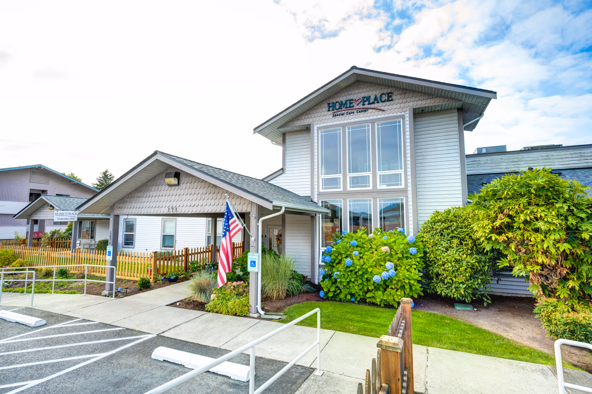 Exterior view of HomePlace Special Care at Oak Harbor facility showing a light gray building with large windows, an American flag near the entrance, a wooden fence, and well-maintained landscaping including green bushes and blue flowers under a partly cloudy sky.