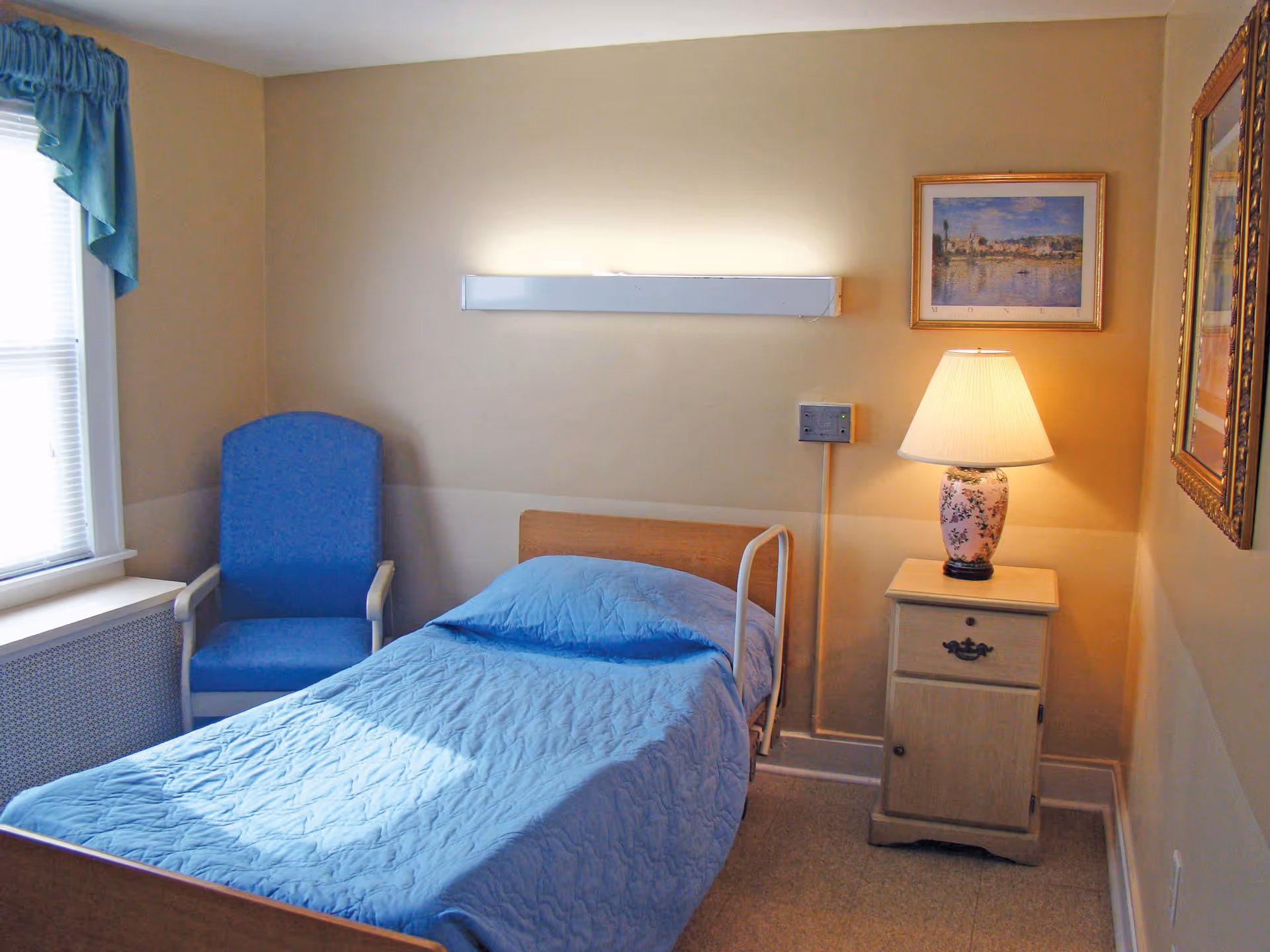 A small nursing center bedroom with a single bed covered in a blue quilt, a blue upholstered chair next to a window with blue curtains, a wooden nightstand with a floral lamp, and framed artwork on the beige walls.