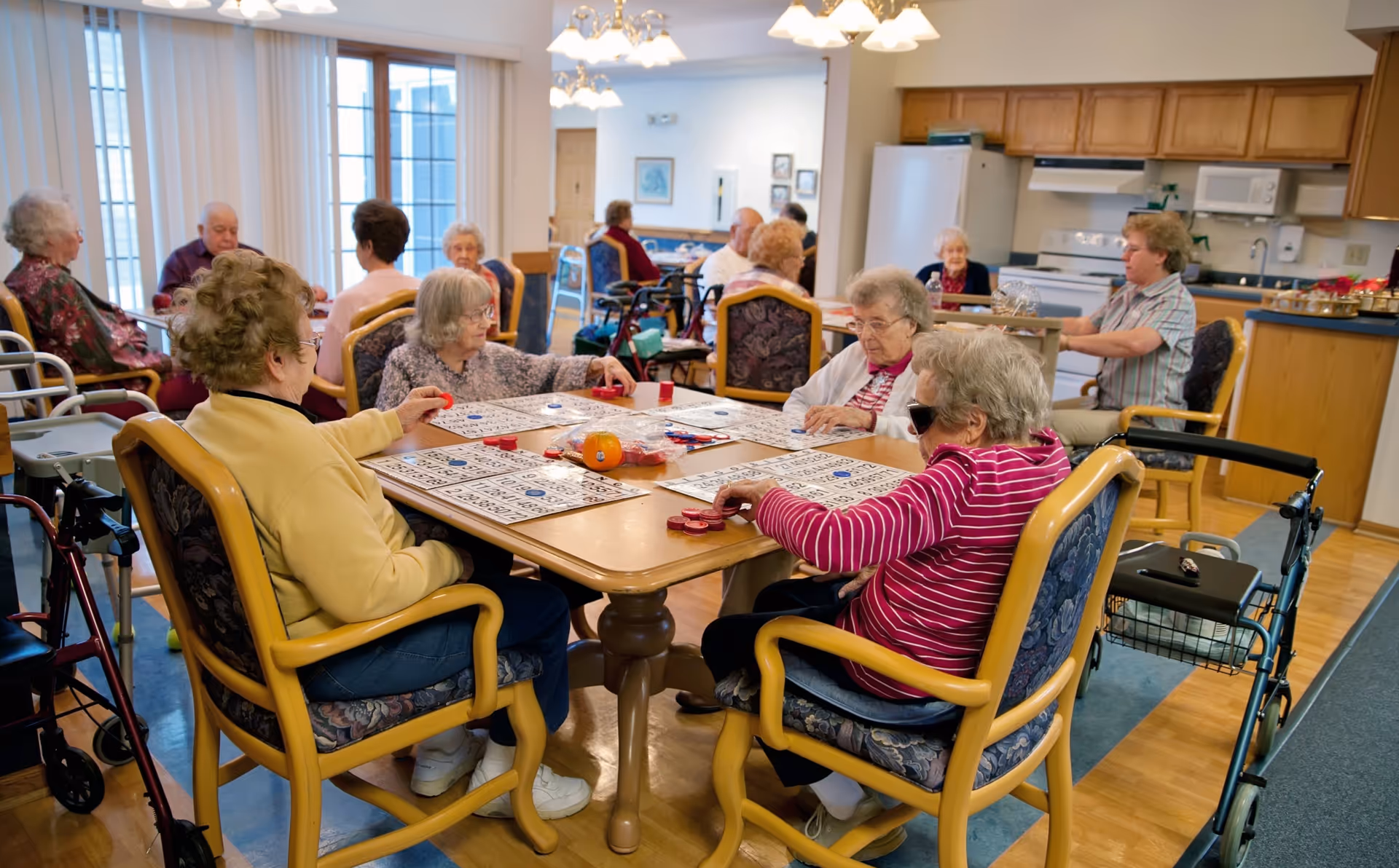 A group of elderly people sitting around a table playing bingo in a well-lit room with large windows and a kitchen area in the background. Several walkers are visible near the chairs, and the participants appear engaged in the game.