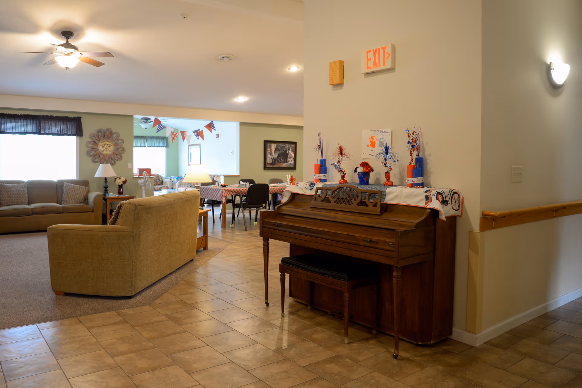 Interior view of a senior living facility common area featuring a wooden piano decorated with patriotic-themed items, a beige couch and loveseat, a ceiling fan, and a dining area with tables and chairs in the background. The floor is tiled near the piano and carpeted near the seating area. An exit sign is visible above the piano.