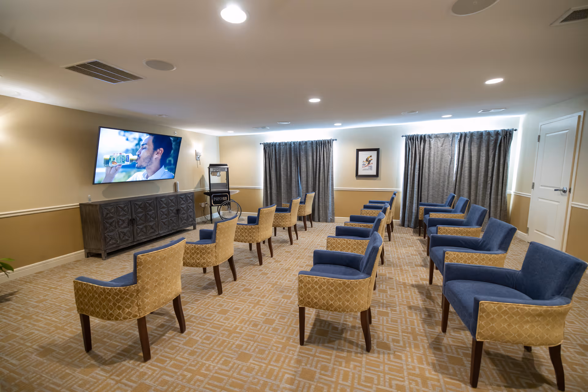 A small theater room with two rows of blue and beige upholstered chairs facing a wall-mounted flat screen TV. The room has beige walls, patterned carpet, gray curtains covering windows, a popcorn machine, and a framed picture on the wall.