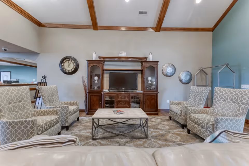 A cozy living room area with four patterned armchairs arranged around a rectangular coffee table. A large wooden entertainment center with a TV and decorative items is against the wall. The room features a high ceiling with wooden beams, a wall clock, and decorative wall hangings.