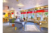 Bright senior living lounge with chairs, small tables, potted plants, and large windows with red valances.