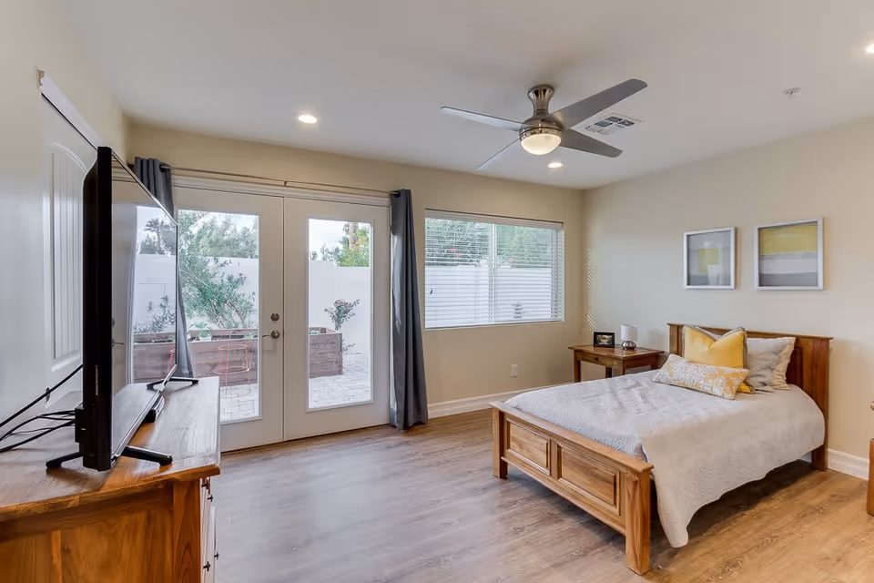 Bright bedroom with a wooden bed, TV on a dresser, ceiling fan, and glass French doors opening to a small patio.