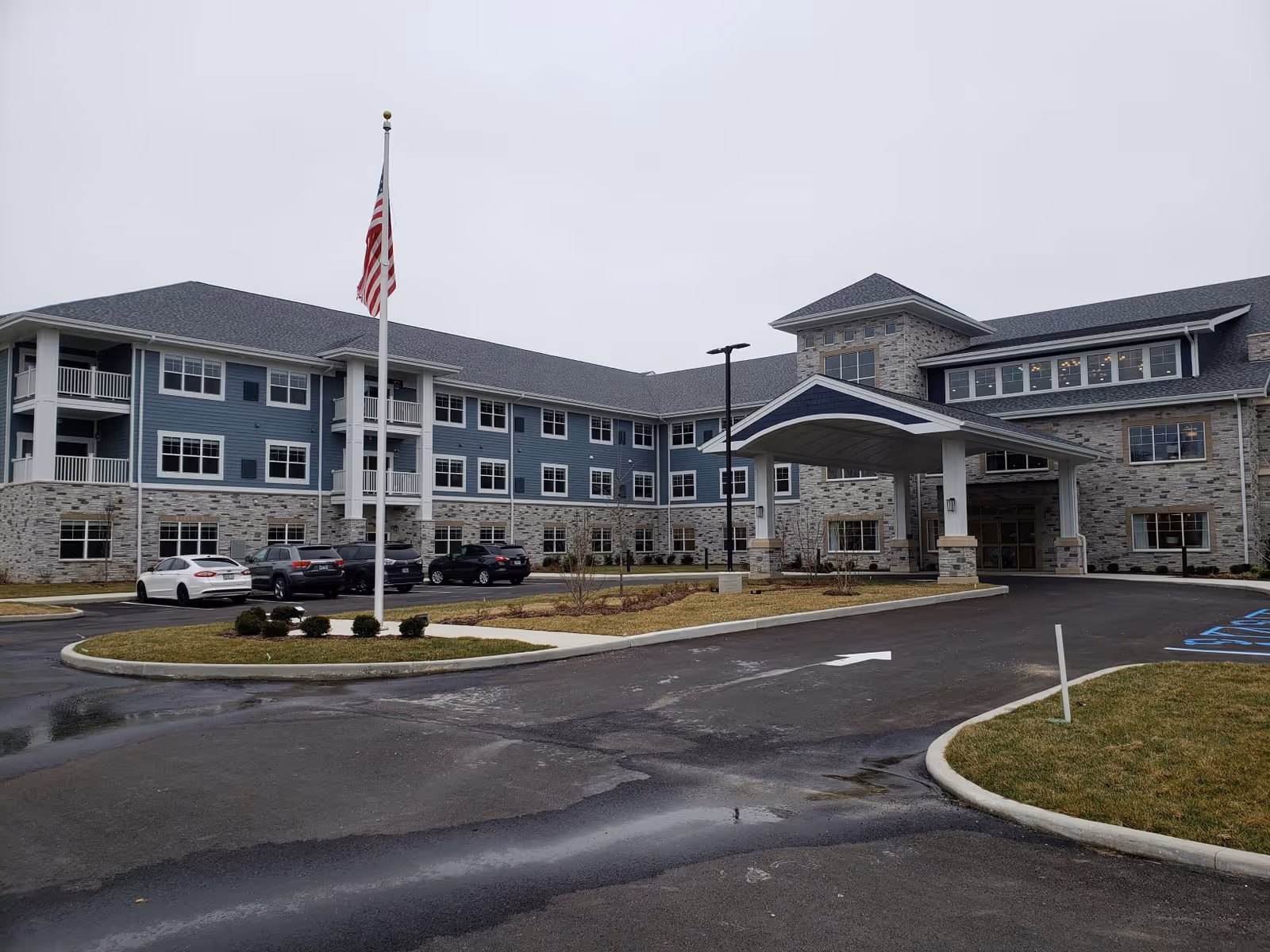 Exterior view of a large senior living facility building with a covered entrance, multiple windows, and a flagpole with an American flag in front. Several cars are parked in the parking lot on a cloudy day.