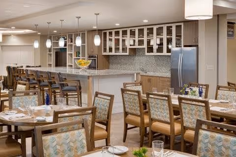 Spacious dining room with multiple set tables and chairs facing a large serving counter and kitchen area with pendant lights and a refrigerator.