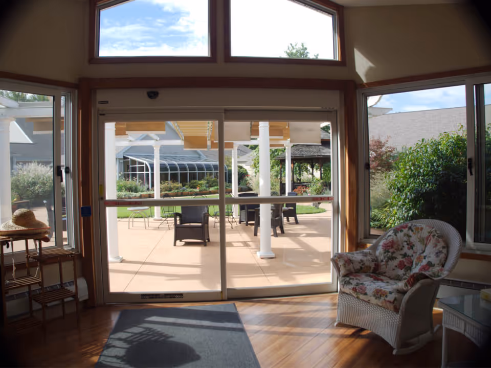 Interior view of a room with large sliding glass doors leading to an outdoor patio area. The room has wooden floors, a floral cushioned wicker chair, a small table with a straw hat, and large windows letting in natural light. Outside, there are chairs and tables on the patio with greenery and buildings in the background under a partly cloudy sky.
