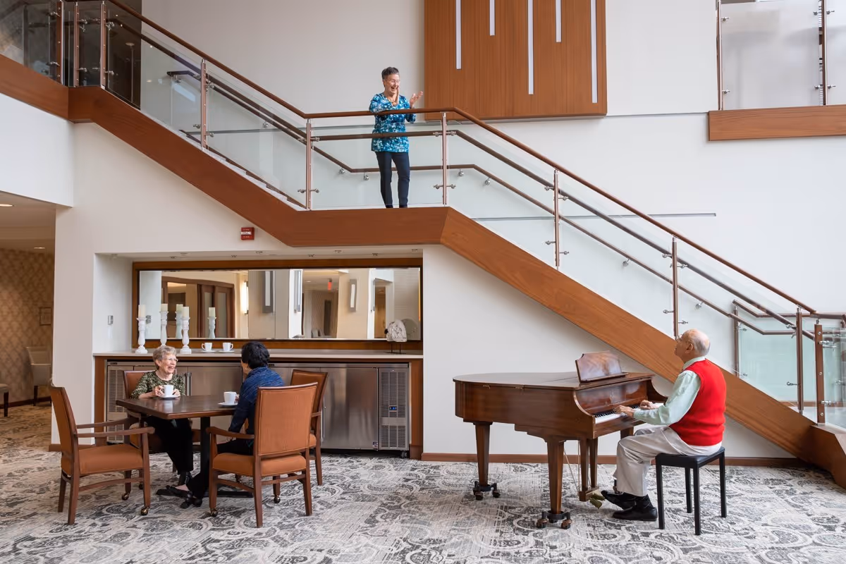 A senior man plays a grand piano in a bright atrium while two women sit at a nearby table and another woman stands on the upper landing.