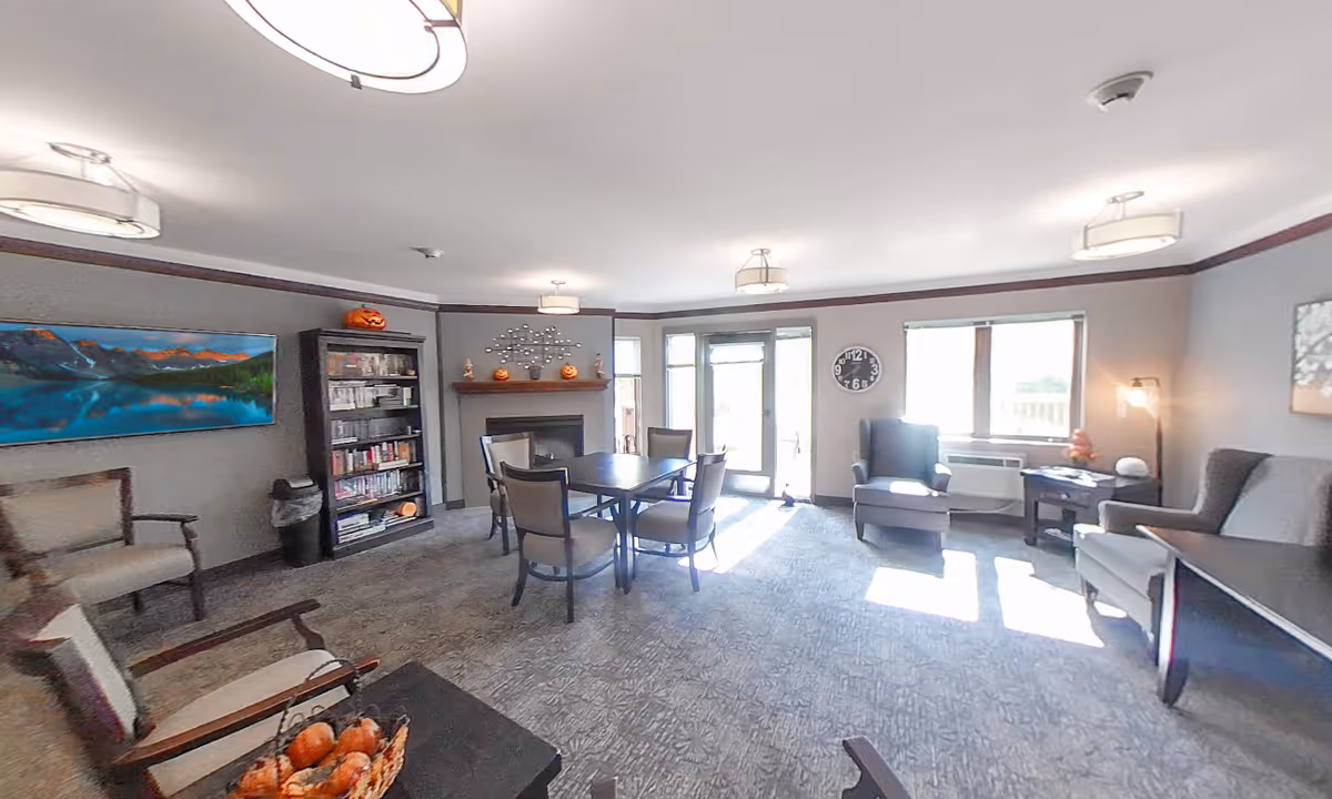 Communal living room with chairs, a central table, bookshelf, fireplace decorated with pumpkins and large sunlit windows.