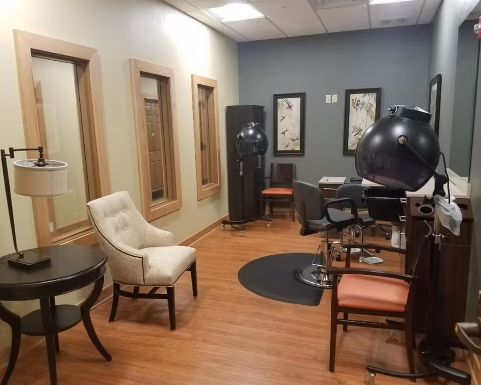 Interior view of a salon area in a senior living facility with wooden flooring, a beige upholstered chair next to a round side table with a lamp, two black hair drying stations with chairs, and framed artwork on the walls.