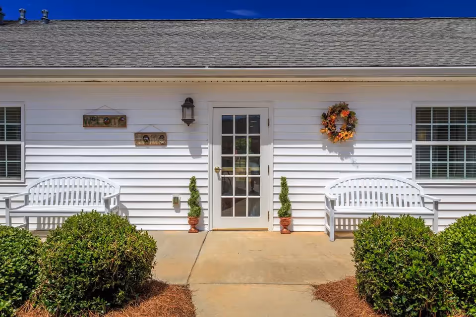 Front entrance of a white single-story building with a glass door flanked by two white benches, decorative wreath and potted shrubs.