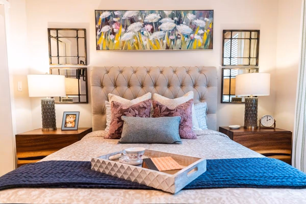 A neatly made bedroom with a tufted headboard, decorative pillows, bedside tables with lamps and mirrored wall art, and a tray on the bed.