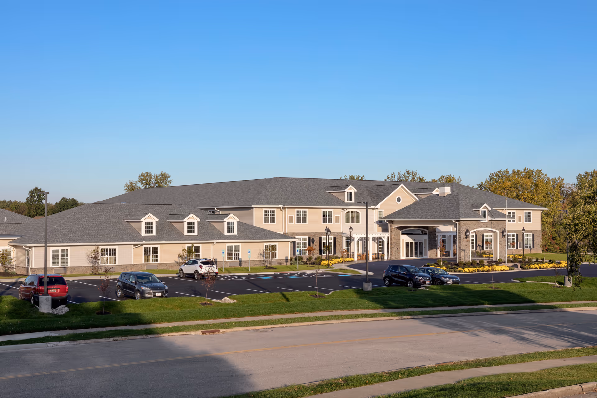Front exterior of a two-story assisted living building with a covered entrance, parking lot, and landscaped lawn under a clear blue sky.