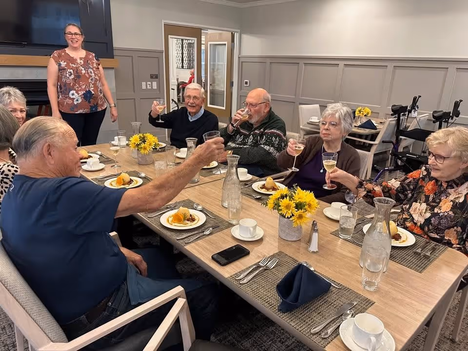 A group of elderly people sitting around a dining table in a senior living facility, raising their glasses in a toast. The table is set with plates of dessert, cups, and water pitchers, and decorated with small vases of yellow flowers. A woman stands smiling in the background near a fireplace and television.