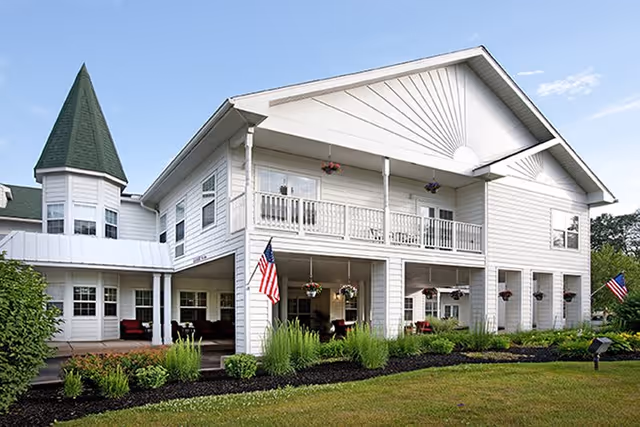 Exterior view of a two-story white building with a green roof and a turret. The building has a covered porch with hanging flower baskets and American flags displayed. There is a well-maintained lawn and garden beds in front of the building under a clear blue sky.