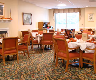 A dining room with multiple tables covered in white tablecloths and set with dining ware. The room has wooden chairs with red cushions, a patterned carpet, a fireplace on the left wall, and a large window with curtains letting in natural light.