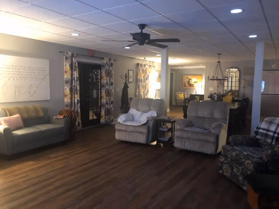 A cozy living room area in Magnolia Assisted Living featuring a gray sofa with a pink pillow and a brown throw, two beige recliners with a small side table between them, and a floral patterned armchair. The room has wood flooring, a ceiling fan, recessed lighting, and floral curtains on a glass door. In the background, there is a dining area with a chandelier and wall decorations.