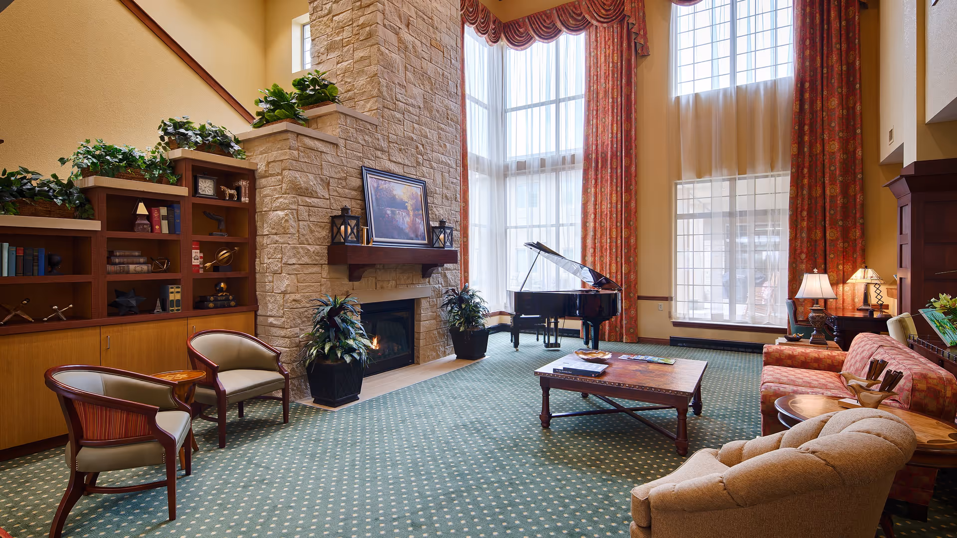 A spacious and elegant living room with high ceilings and large windows covered by red patterned curtains. The room features a stone fireplace with a painting and lanterns on the mantel, a black grand piano near the windows, a green carpet with a dotted pattern, a wooden coffee table, a red patterned sofa, beige armchairs, side tables with lamps, and built-in wooden shelves with books and decorative items.