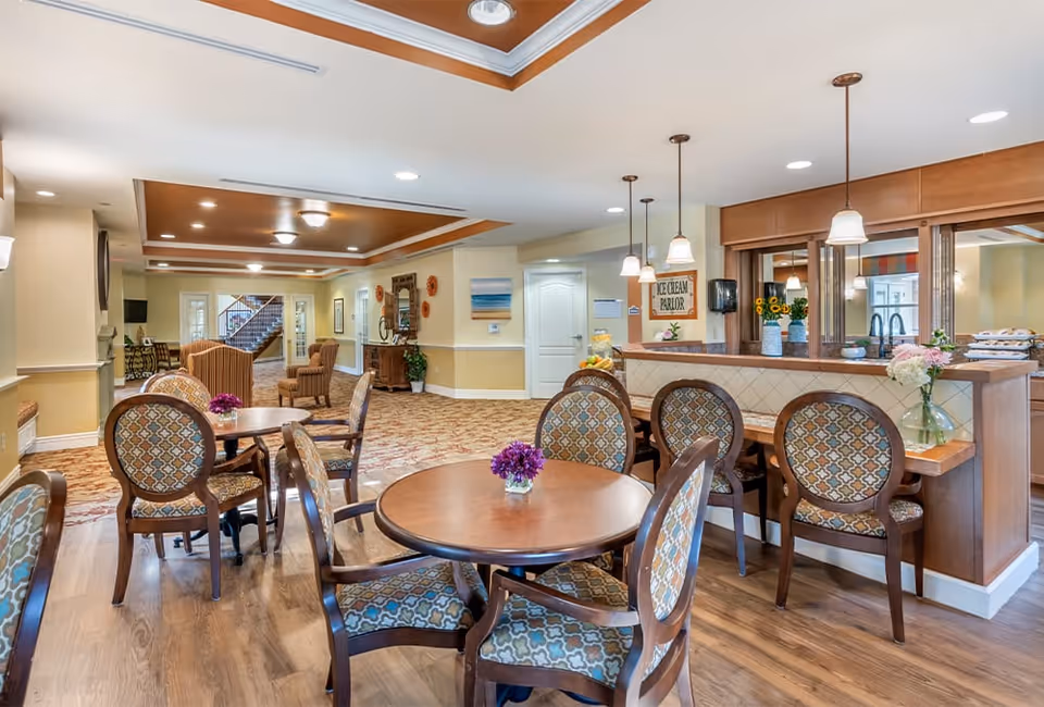 Interior view of a senior living facility common area with round wooden tables and patterned chairs. There are flowers in small vases on the tables. To the right, there is a counter with bar stools and pendant lights hanging above. The area has warm lighting, wooden flooring, and a carpeted section leading to a hallway with more seating and a staircase in the background.