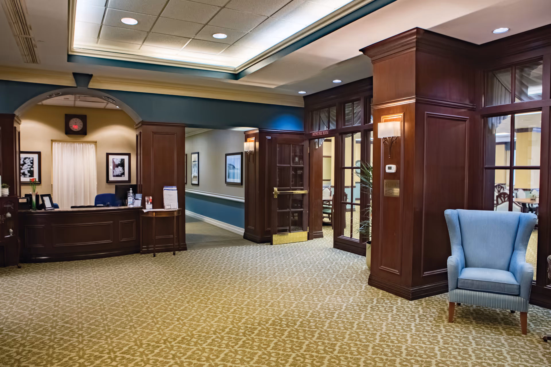 Interior view of a senior living facility lobby area with a reception desk on the left, a blue upholstered armchair on the right, and a hallway leading to other rooms. The space features patterned carpet, wood paneling, framed artwork on the walls, and soft ceiling lighting.