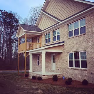 Exterior view of a two-story brick building with multiple windows, a covered porch supported by white columns, and a small balcony on the upper floor. The building is surrounded by a lawn and trees in the background under a clear sky.