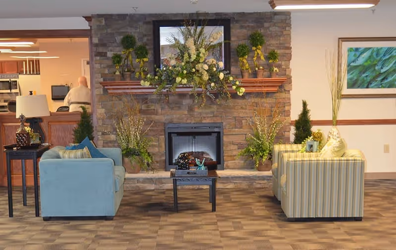 A cozy living room area with a stone fireplace decorated with floral arrangements and a framed picture above it. Two armchairs, one blue and one striped, face each other with a small black coffee table in between. There is a side table with a lamp and decorative items on the left, and a framed green artwork on the wall to the right. The room has carpeted flooring and soft lighting.