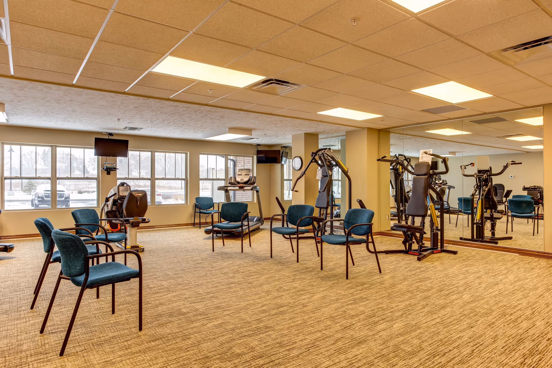 A fitness room in a senior living facility with exercise equipment including a treadmill, stationary bike, and weight machines. Several teal upholstered chairs are arranged in the center of the room. Large windows let in natural light and a wall-length mirror reflects the equipment and chairs.