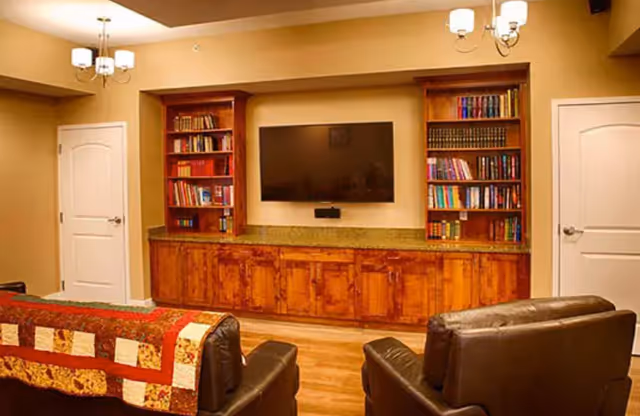 A cozy living room with two brown leather armchairs and a couch covered with a patchwork quilt facing a wall-mounted flat screen TV. The TV is flanked by two wooden bookshelves filled with books, and below the TV is a long wooden cabinet with multiple doors. The room has warm beige walls, wooden flooring, two white doors on either side of the TV, and two ceiling light fixtures with multiple bulbs.