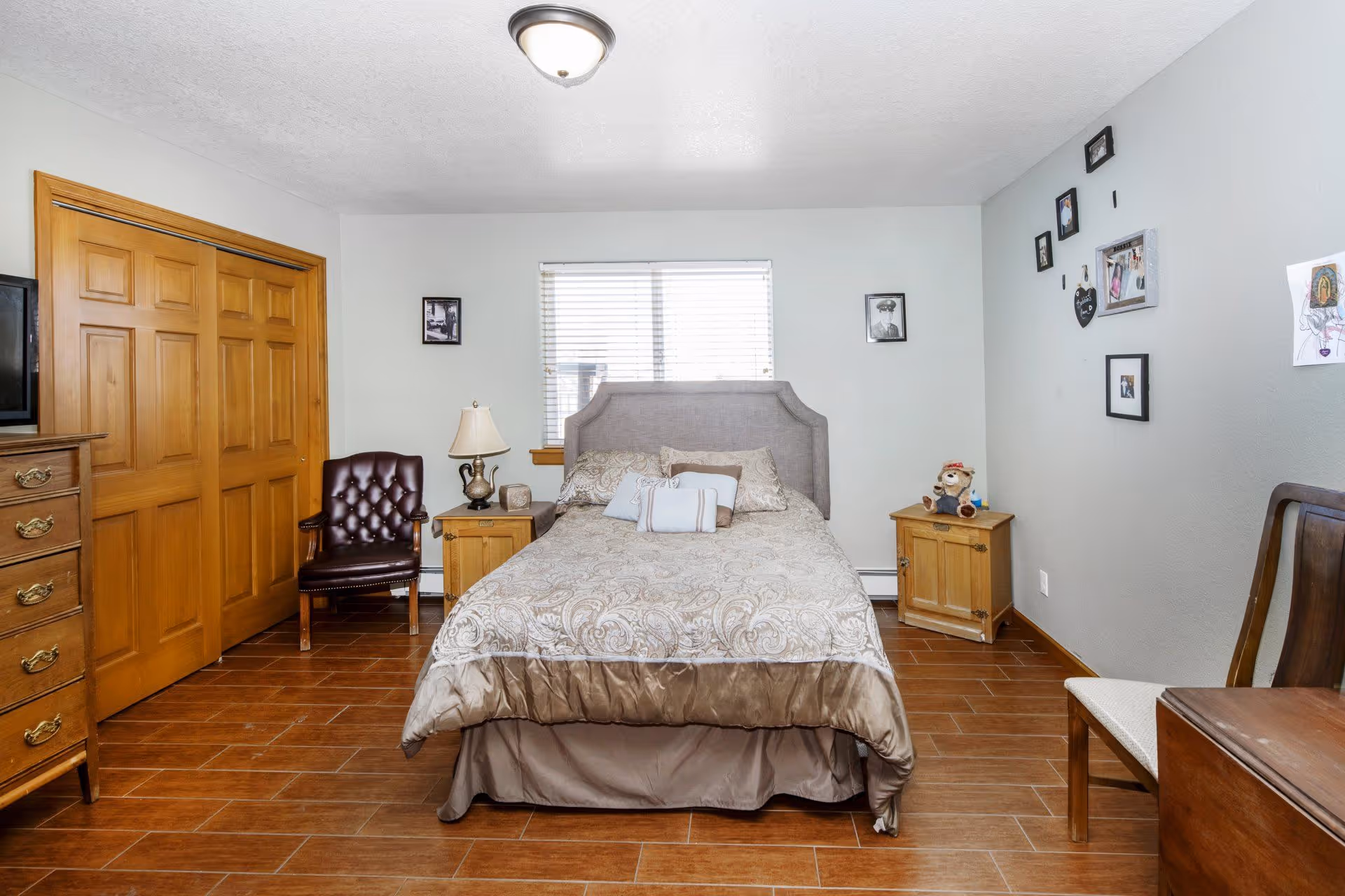 Bedroom with a double bed centered under a window, wooden nightstands and dresser, a leather chair, and framed photos on the walls.