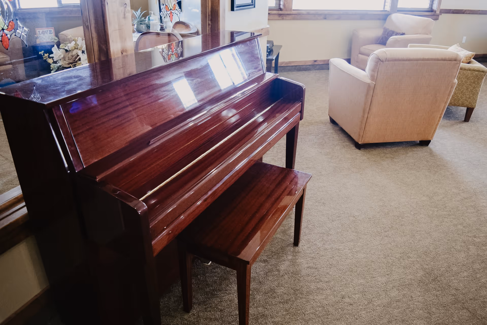 A polished wooden upright piano with a matching bench in a carpeted room. In the background, there are beige upholstered armchairs and a small table near windows with wooden trim. A decorative glass panel with stained glass accents is also visible behind the piano.