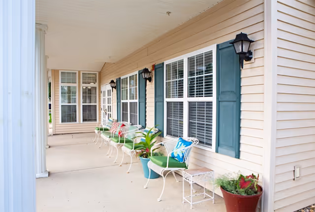 Covered outdoor porch area with white metal chairs featuring green cushions and colorful pillows, potted plants, beige siding with blue shutters, and black wall-mounted lanterns.