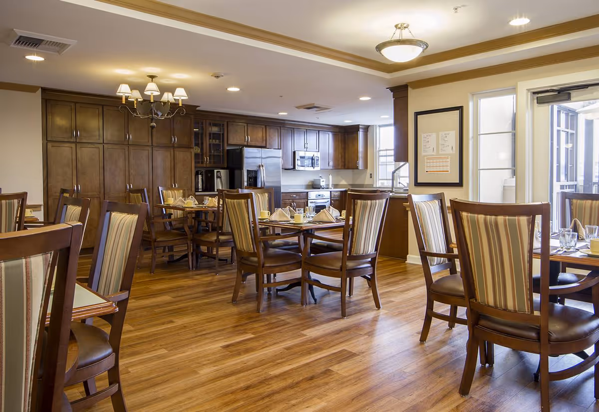 A dining area with several wooden tables and chairs featuring striped upholstery. The room has wooden flooring and a kitchen area with dark wood cabinets, stainless steel appliances including a refrigerator, microwave, and oven. The space is well-lit with ceiling lights and a chandelier, and there is a door leading outside.