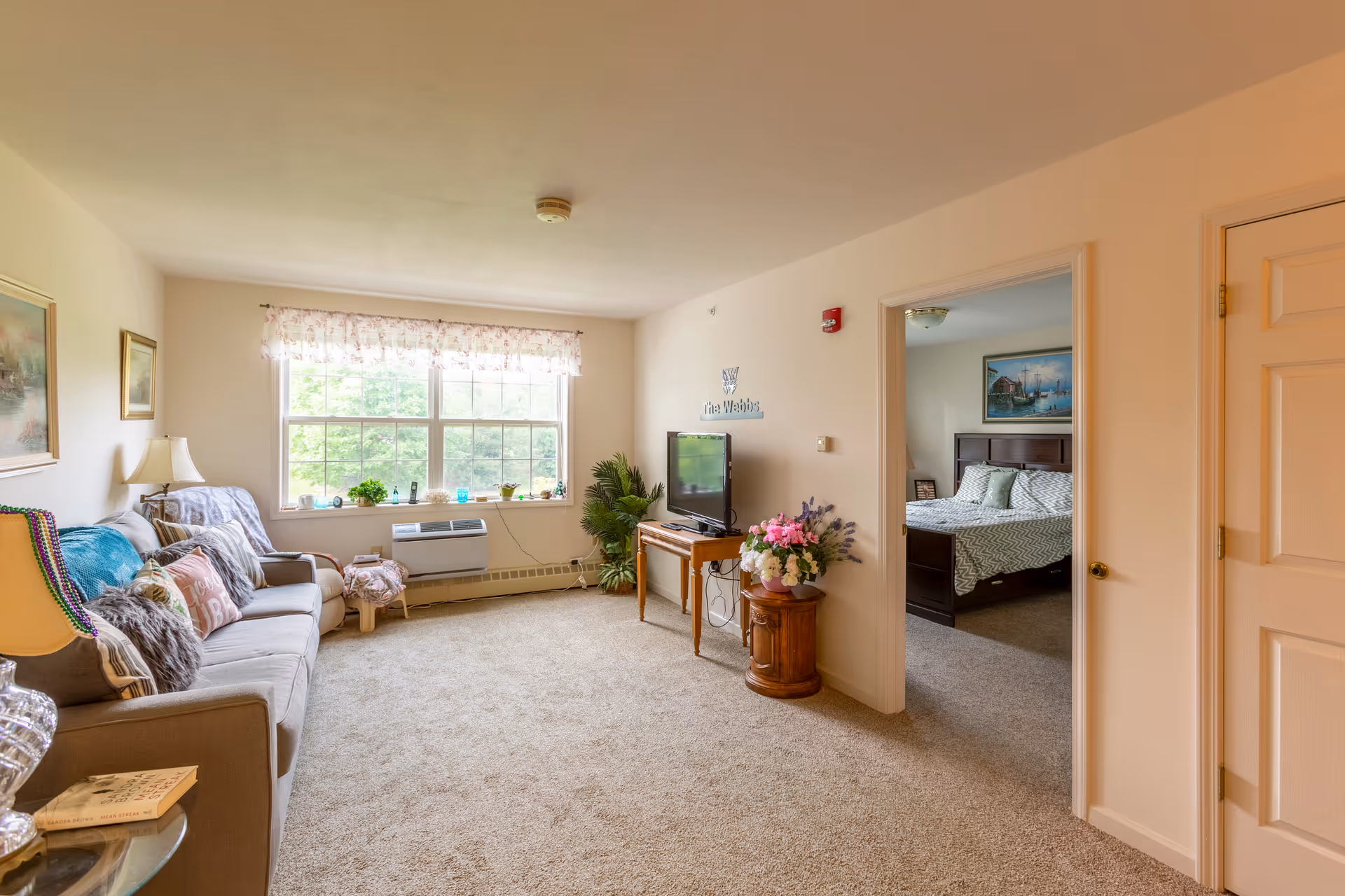 A cozy living room with a beige carpet, a gray sofa adorned with colorful pillows and a throw blanket, a side table with a lamp and a book, a large window with floral curtains letting in natural light, a TV on a wooden stand, and a small round table with a flower arrangement. An open doorway leads to a bedroom with a bed, pillows, and a painting on the wall.