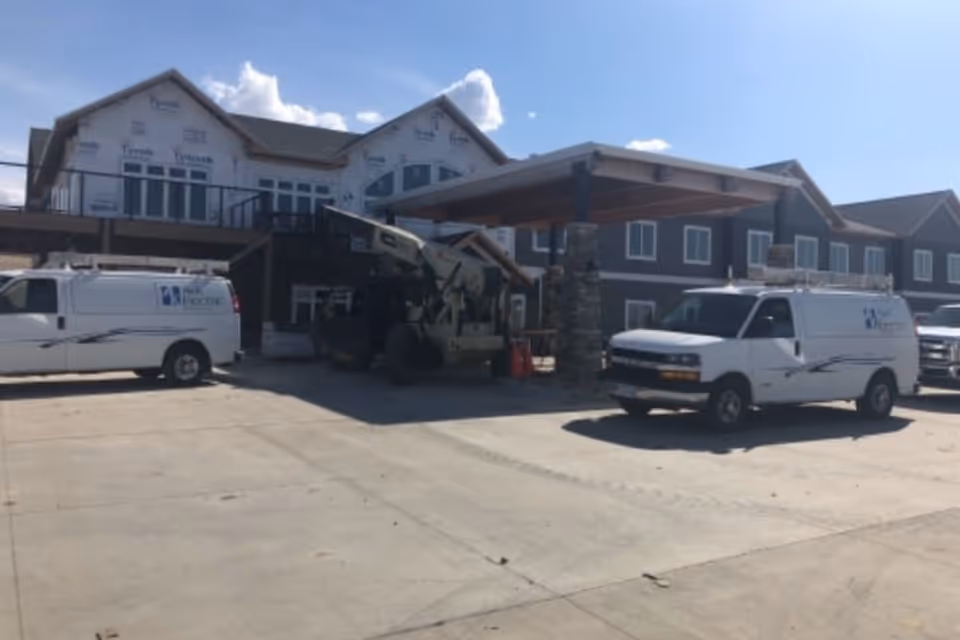 Exterior view of a large building under construction with a covered entrance supported by stone pillars. Two white vans and construction equipment are parked in front on a concrete driveway under a clear blue sky.