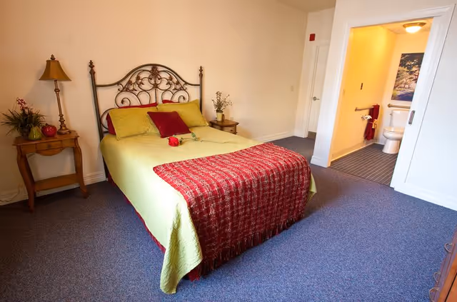 A bedroom with a decorative metal headboard bed covered with a green bedspread and red throw blanket, accompanied by red and green pillows. There are two wooden nightstands on either side of the bed, one with a lamp and the other with a small plant. An open door reveals a bathroom with a toilet, towel rack, and a framed picture on the wall.