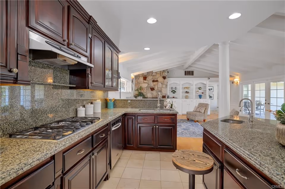 A spacious kitchen with dark wood cabinets and granite countertops. The kitchen features a gas stove, dishwasher, and two sinks. In the background, there is a living area with a stone fireplace, built-in white shelves, a beige armchair, and large windows letting in natural light.