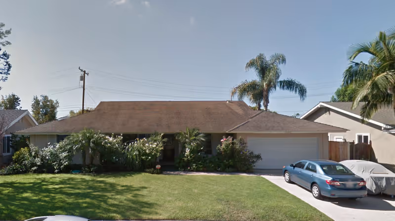 Single-story residential building with a brown shingle roof, surrounded by green shrubs and palm trees. There is a well-maintained lawn in front and a driveway with two parked cars, one covered with a car cover. The sky is clear and blue.