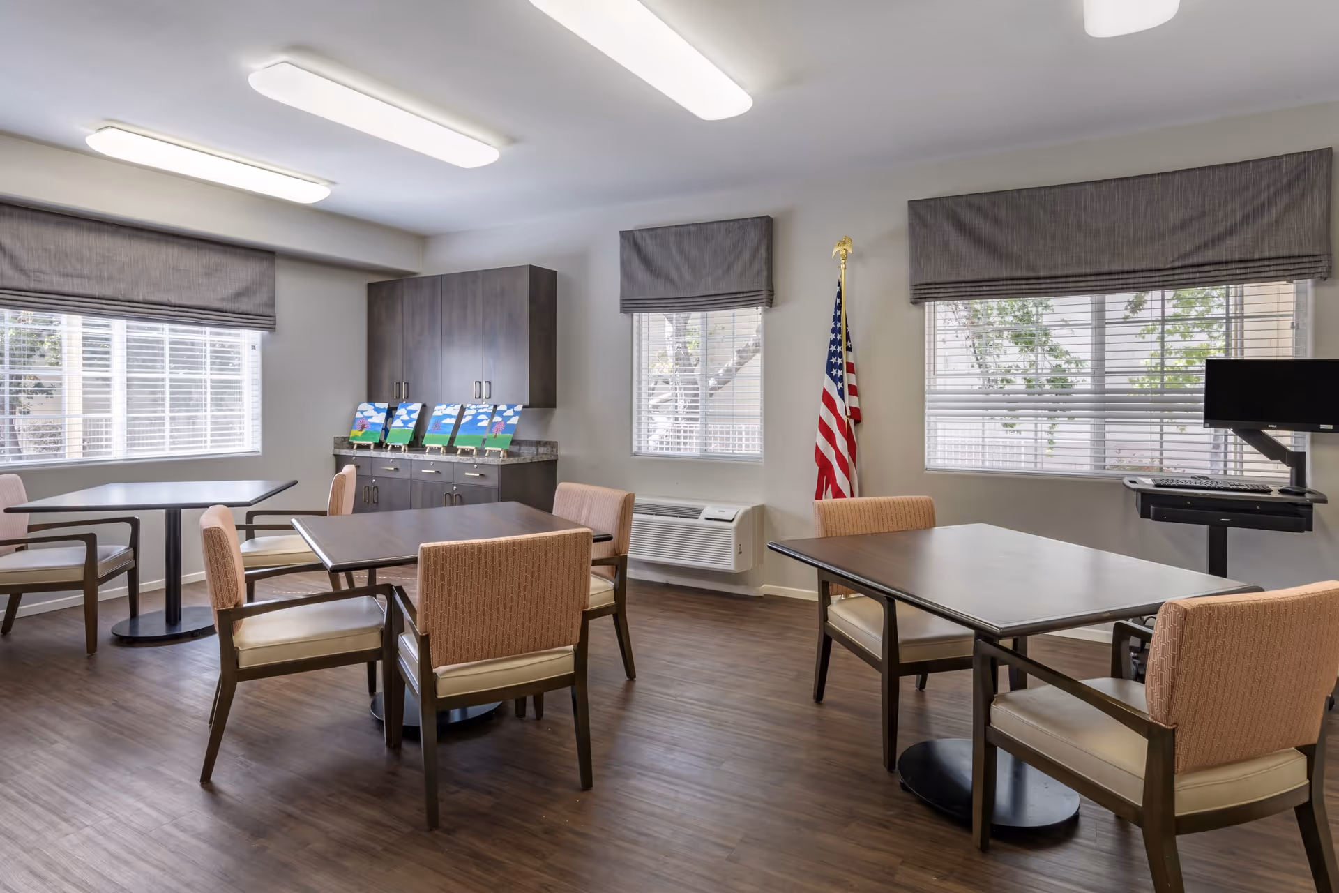 A bright room with three tables, each surrounded by four cushioned chairs. The room has large windows with gray blinds, a cabinet with colorful paintings on top, an American flag, and a computer workstation with a monitor and keyboard on a stand. The floor is wooden and the ceiling has rectangular fluorescent lights.