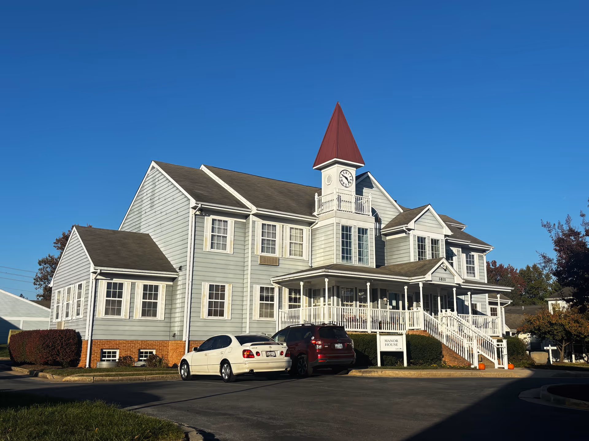 Front view of a light-gray multi-story building with a clock tower, wraparound porch and cars parked in front under a clear blue sky.