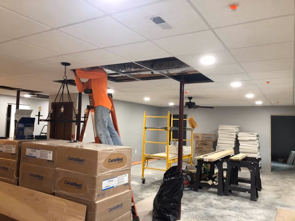 A worker on a ladder installs ceiling tiles in a renovation area with stacked boxes, scaffolding, and construction materials.