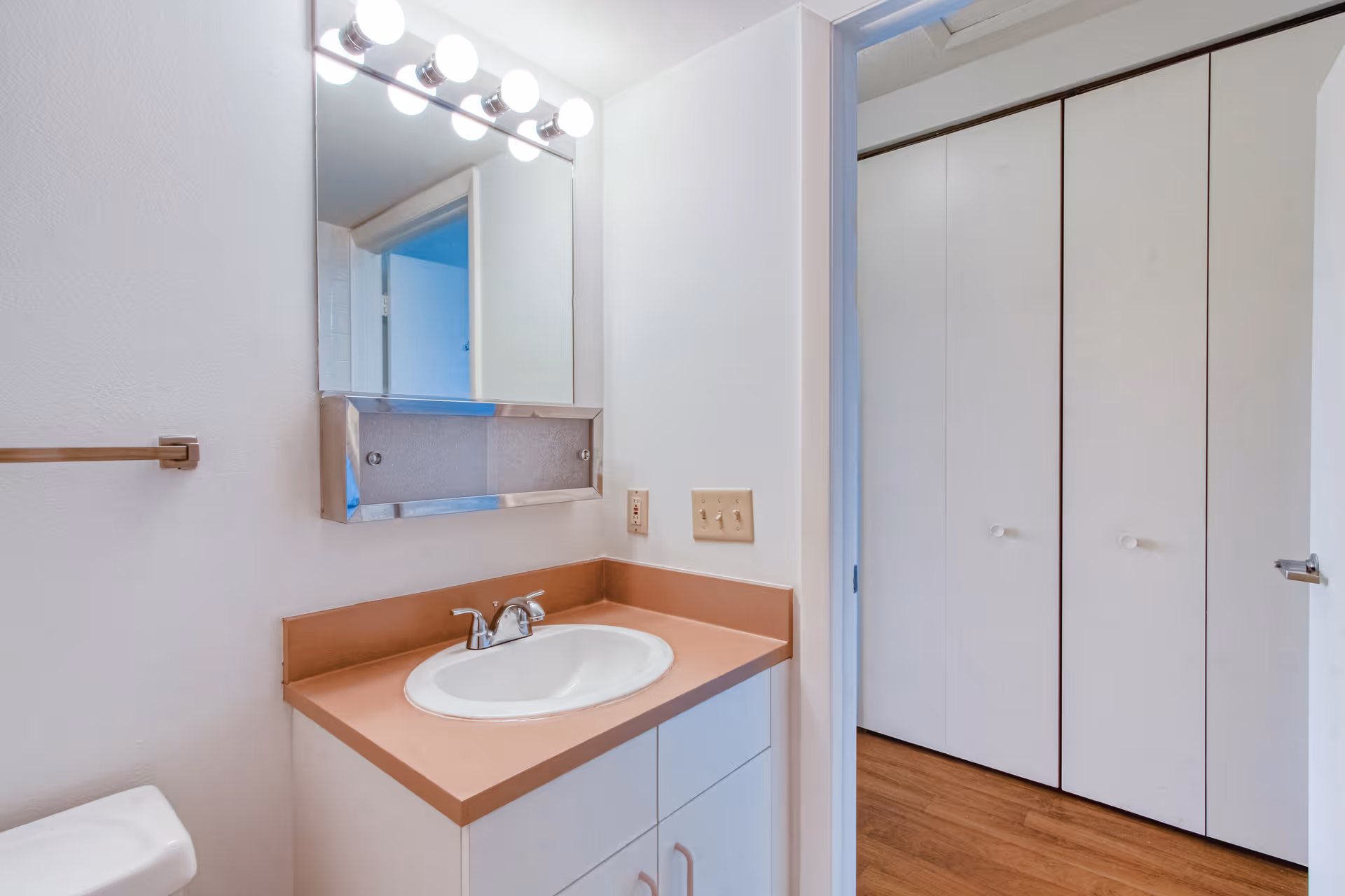 A bathroom vanity with a sink, countertop, and a mirror above it. The mirror has lights on top. To the left, part of a toilet is visible. The bathroom has white walls and a towel rack. Through an open door, a hallway with wooden flooring and white closet doors is visible.