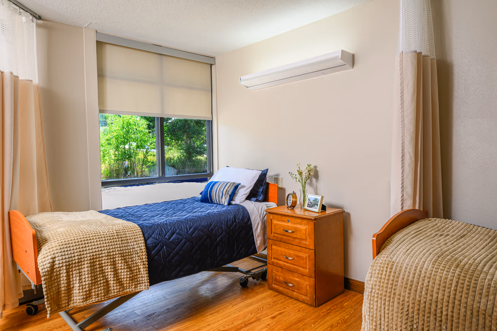 A bright and clean bedroom in a senior living facility with two single beds. One bed has a navy blue quilt and striped pillows, while the other has a beige textured blanket. Between the beds is a wooden nightstand with three drawers, holding a small clock, a framed photo, and a vase with flowers. A large window with a beige roller shade lets in natural light, and the room has light-colored walls and wood flooring.