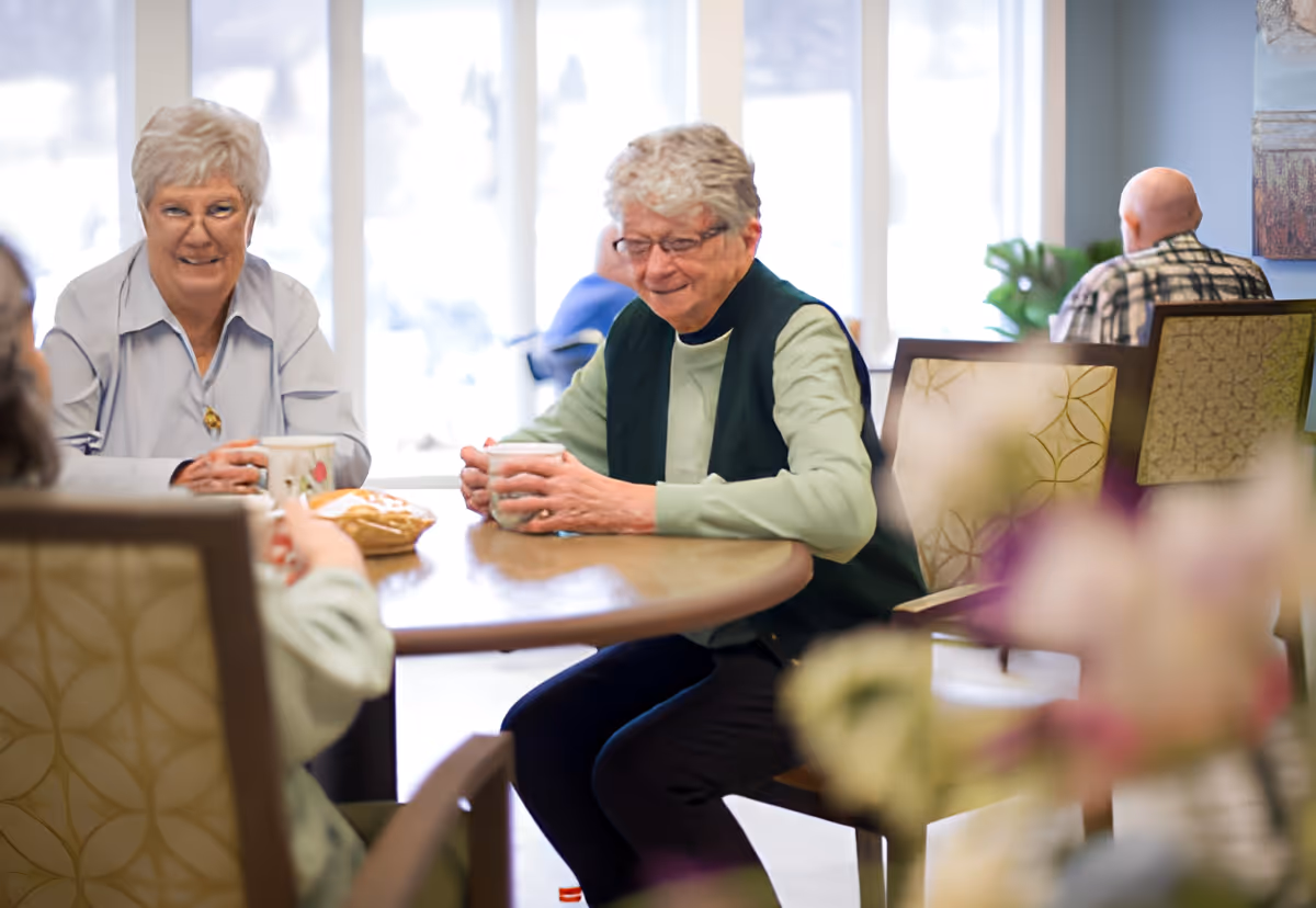 Three elderly people sitting around a round table in a well-lit room, holding mugs and engaging in conversation. The room has large windows letting in natural light and comfortable chairs with patterned upholstery.