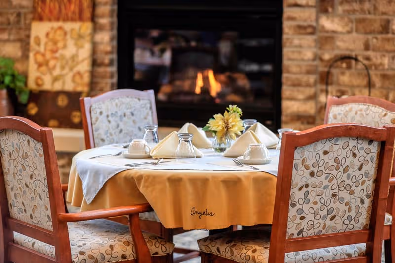 A round dining table set for four with folded napkins, cups, and glasses, surrounded by wooden chairs with patterned upholstery. In the background, there is a lit fireplace with a brick surround and some decorative items on the mantel.