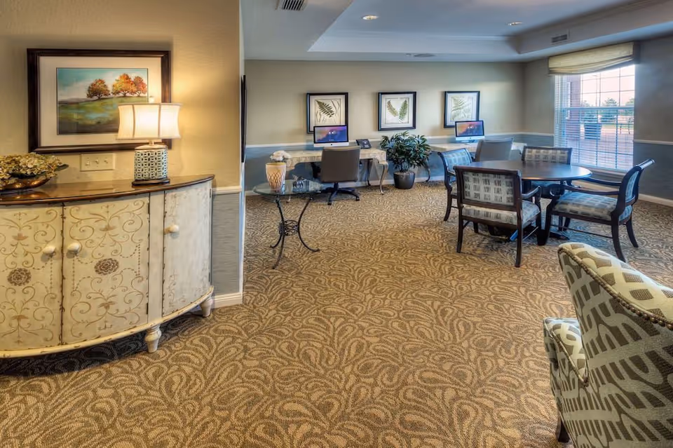 A well-lit interior room with patterned carpet and beige walls. On the left, there is a decorative cabinet with a lamp and a framed picture above it. In the background, there are two computer desks with chairs and framed botanical prints on the wall. To the right, there is a round table with four upholstered chairs near a large window with blinds. A patterned armchair is partially visible in the foreground.