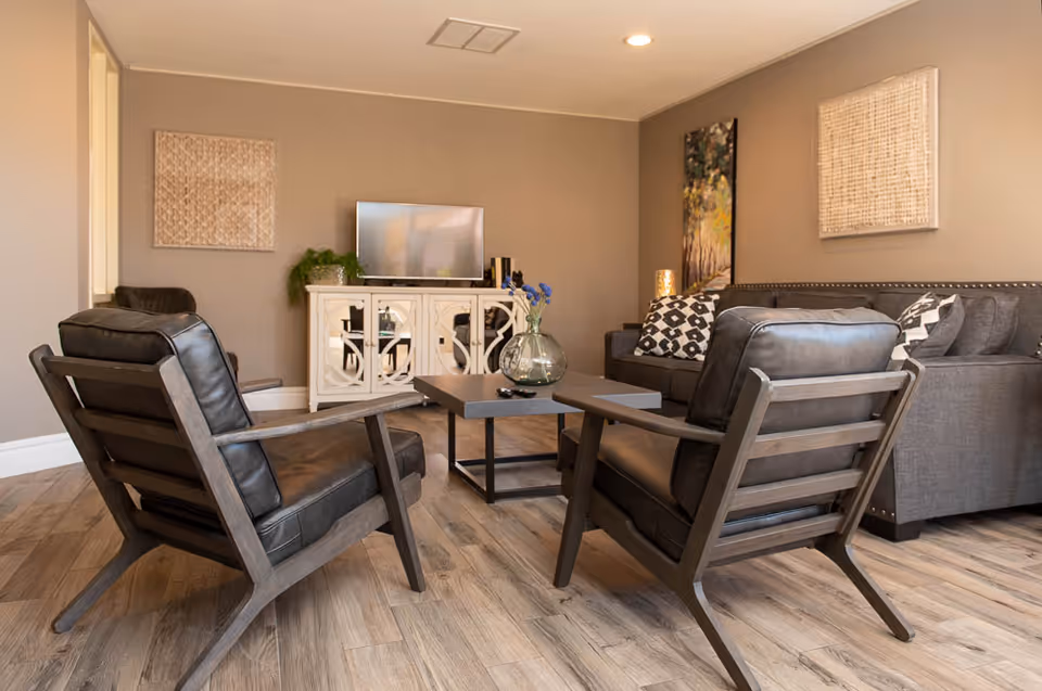 A cozy living room with two dark leather armchairs facing a gray sofa with patterned cushions. A modern coffee table with a glass vase holding blue flowers sits in the center. A white cabinet with a TV on top is against the wall, which is decorated with two textured wall hangings and a vertical painting. The floor is wood, and the walls are painted beige.