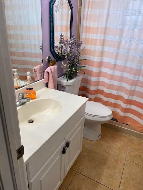 A small bathroom with a white sink and cabinet, a mirror above the sink, a toilet with a vase of purple flowers on the tank, a pink hand towel hanging on a ring, and a shower curtain with horizontal pink and white stripes.
