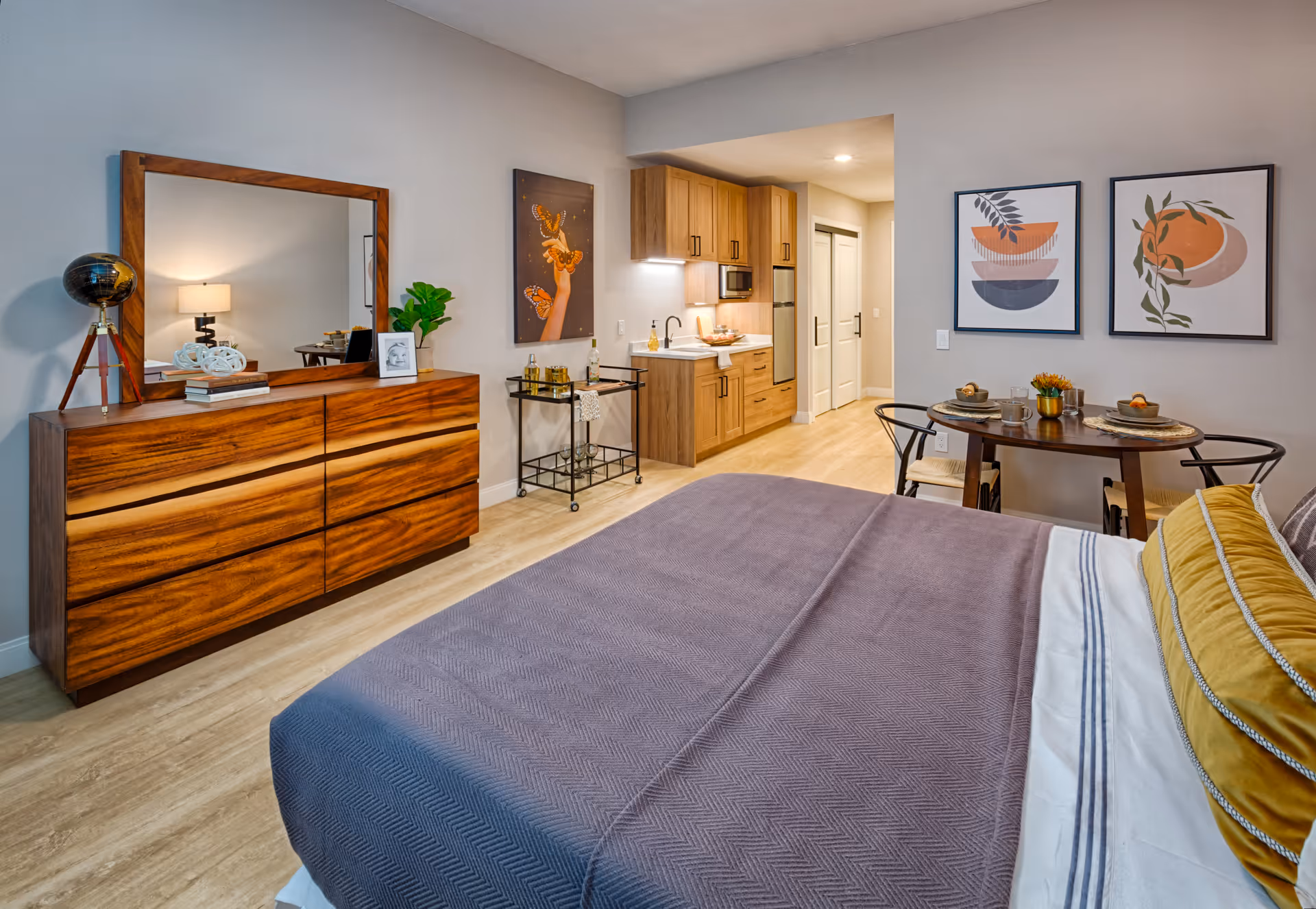 A cozy senior living studio apartment featuring a bed with a purple blanket and yellow pillow, a wooden dresser with a large mirror and decorative items, a small dining table set for two with modern art on the wall, and a compact kitchenette with wooden cabinets and stainless steel appliances.